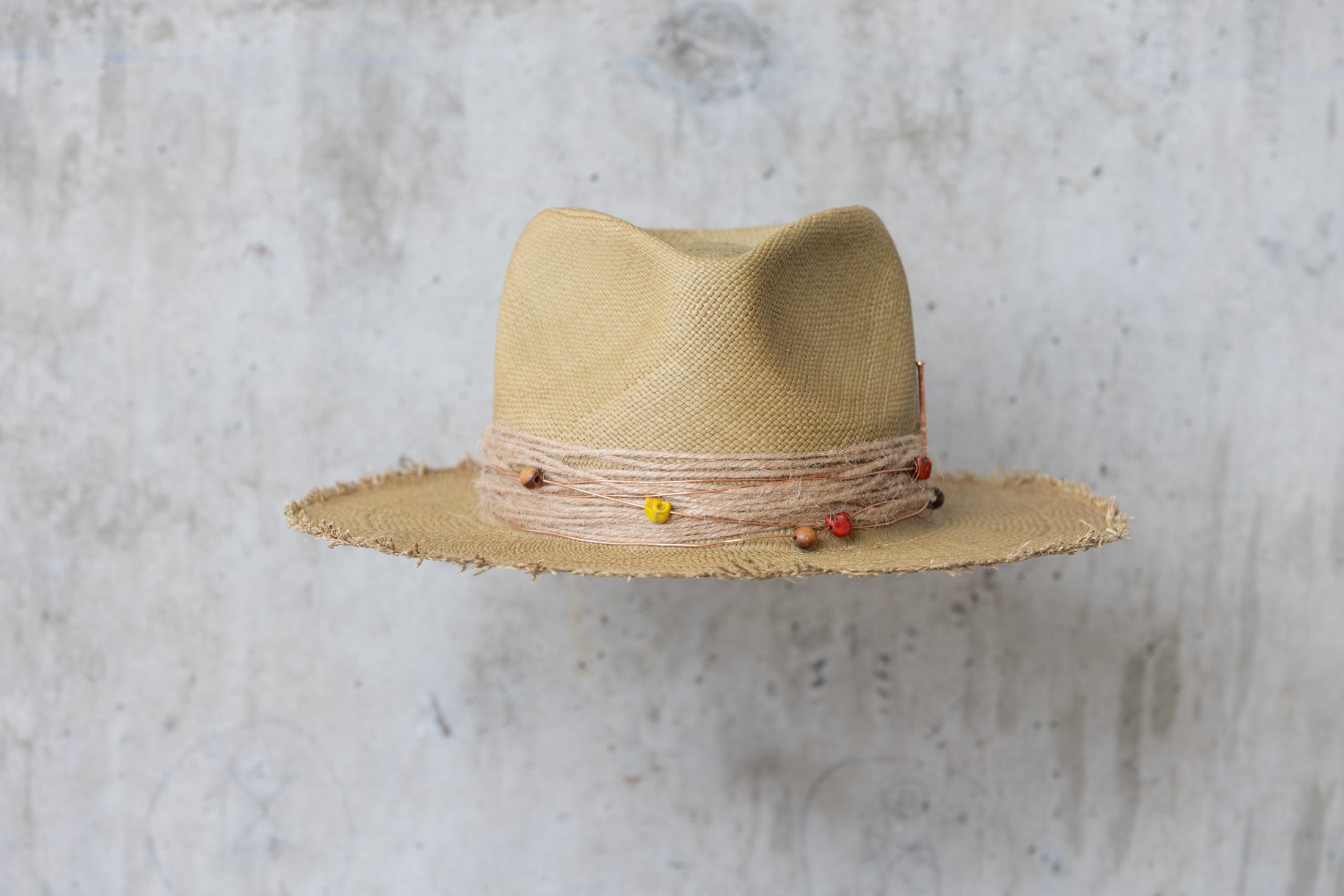 An olive colored straw hat with a teardrop crown and a twine wrap band with copper beads, skull, and wood beads, featuring a frayed edge on the brim front view