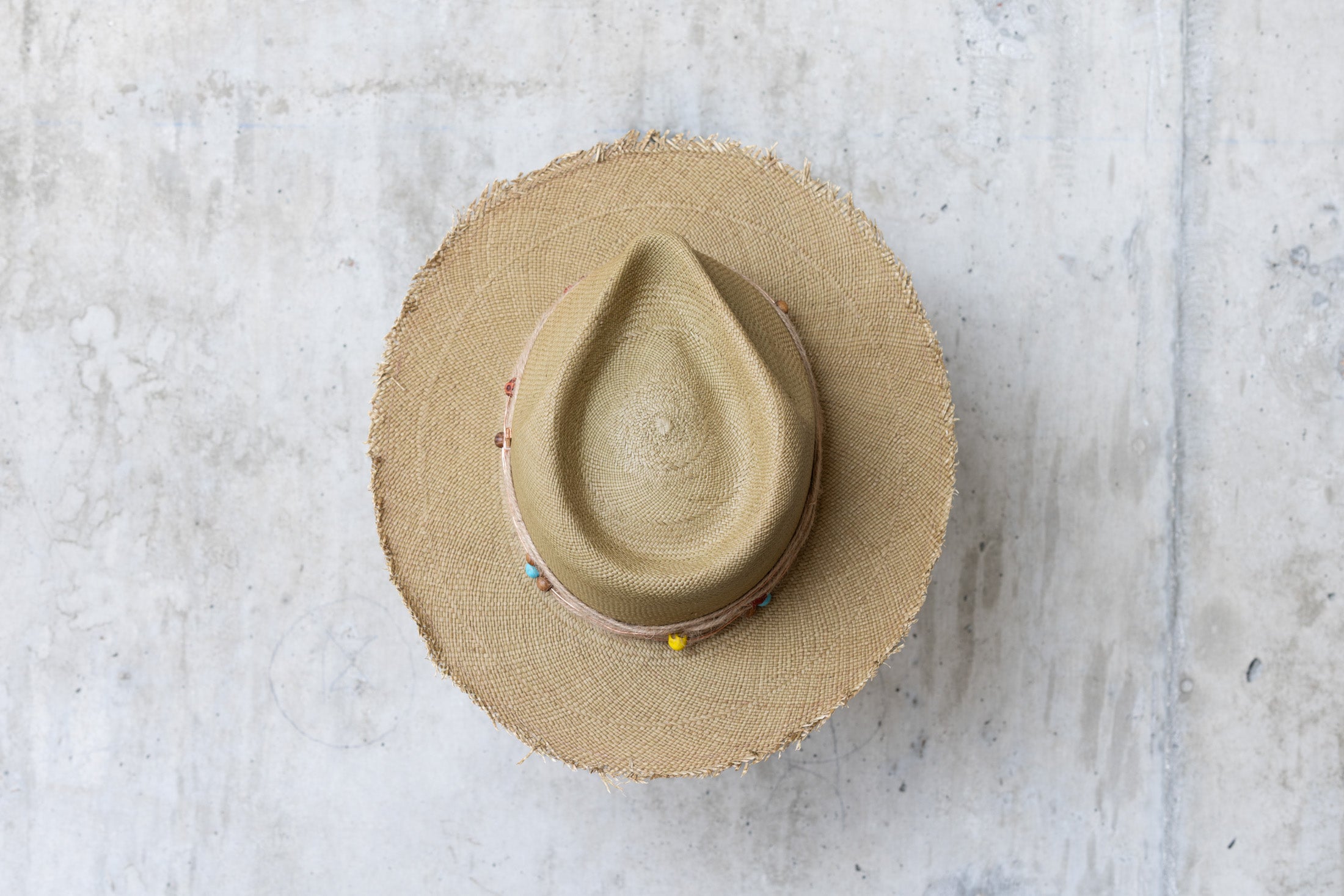 An olive colored straw hat with a teardrop crown and a twine wrap band with copper beads, skull, and wood beads, featuring a frayed edge on the brim top view