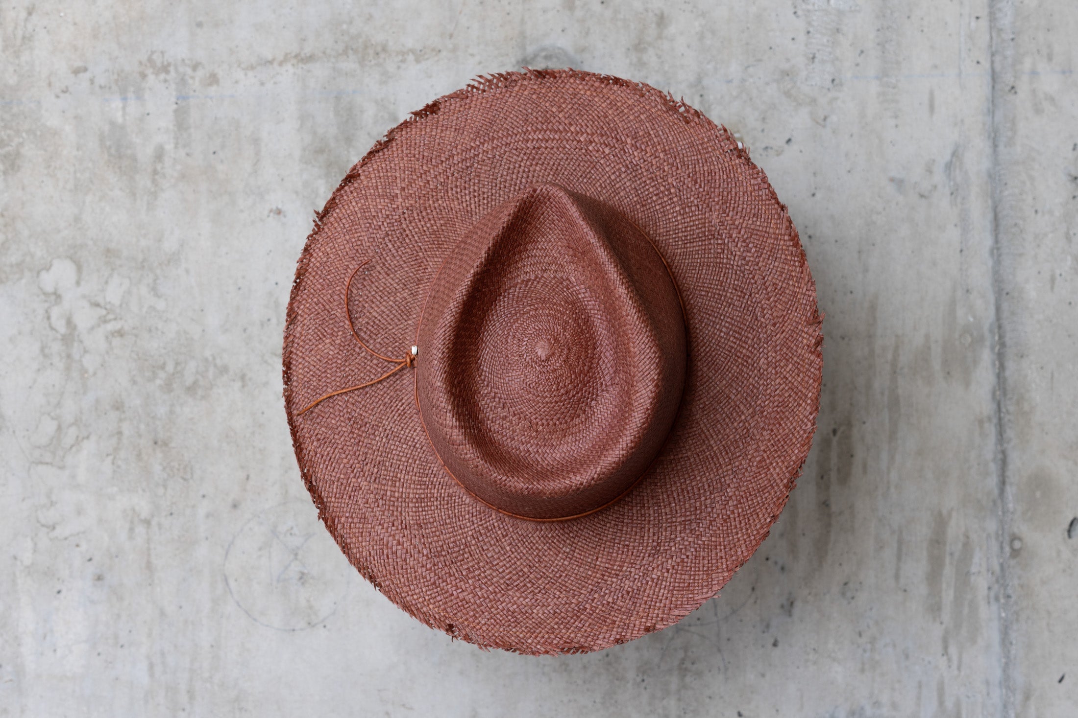 A chocolate brown straw hat with a teardrop crown and a frayed brim edge, with a leather cord wrap top