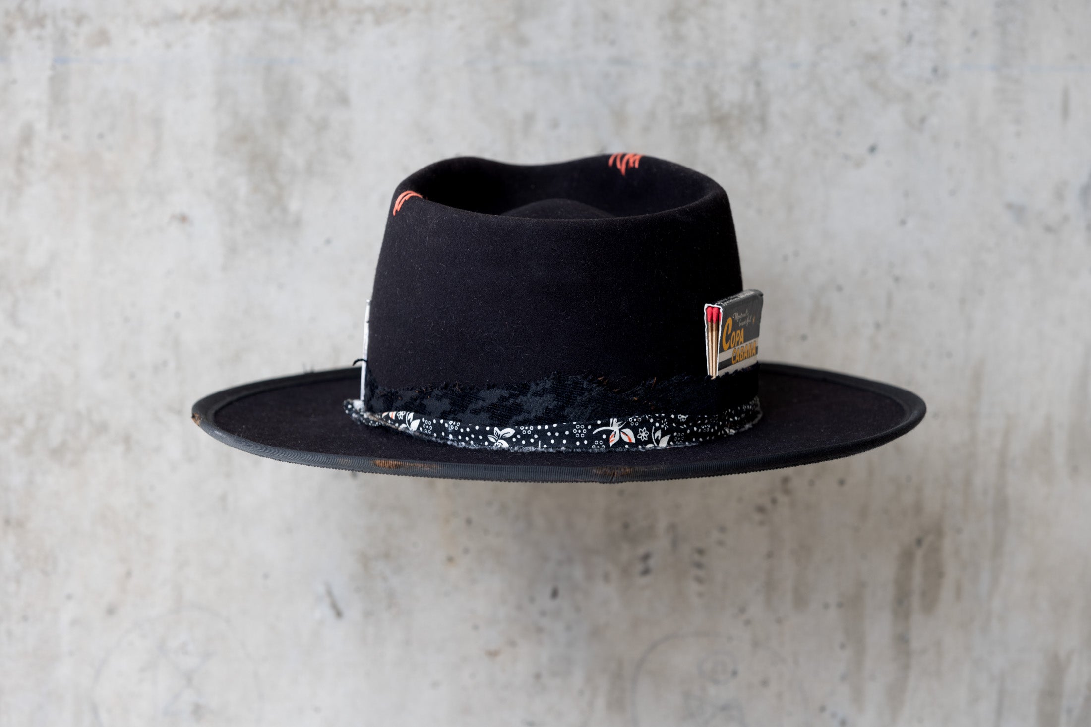 A black fedora western beaver felt hat with a teardrop crown and a brim featuring pencil curls. It has a black houndstooth band with a floral wrap and faded red stitching for men and women back