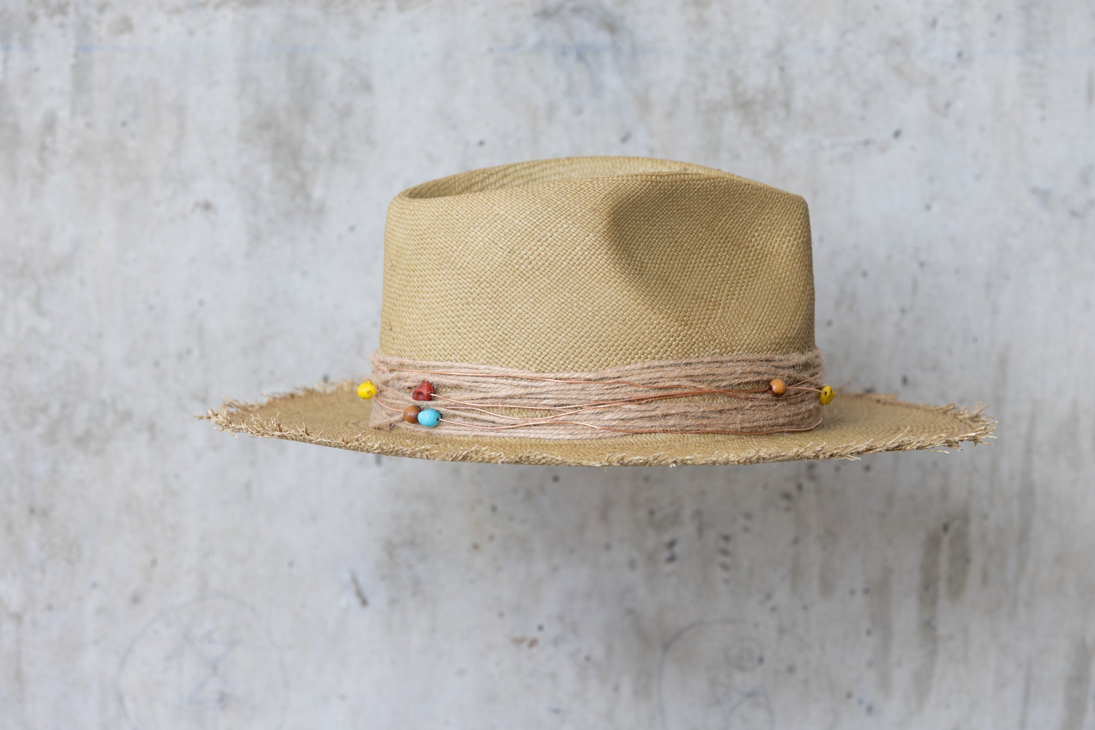 An olive colored straw hat with a teardrop crown and a twine wrap band with copper beads, skull, and wood beads, featuring a frayed edge on the brim right side view