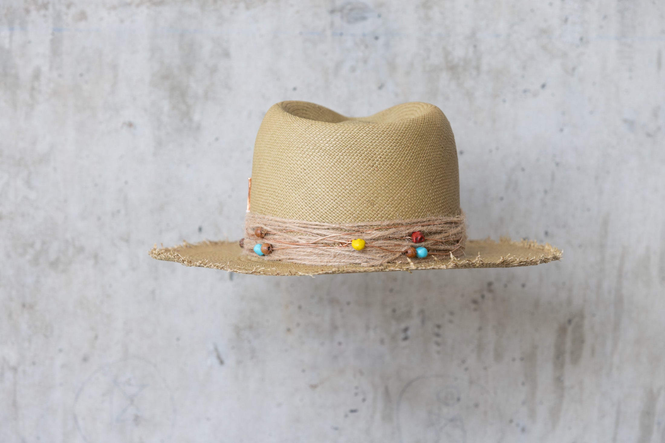An olive colored straw hat with a teardrop crown and a twine wrap band with copper beads, skull, and wood beads, featuring a frayed edge on the brim back side view