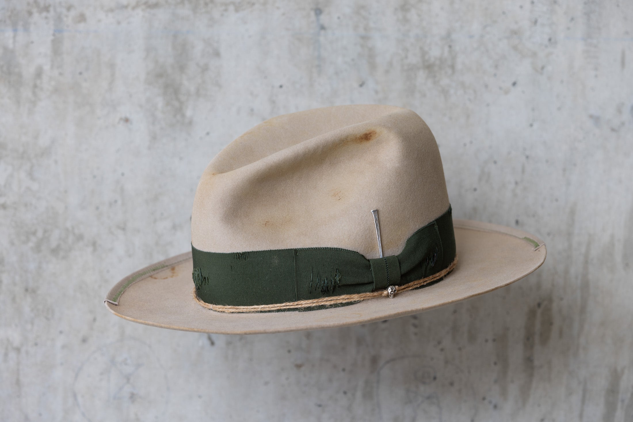 A beige cowboy hat with a forest green grosgrain band and a silver nail on the side angle view
