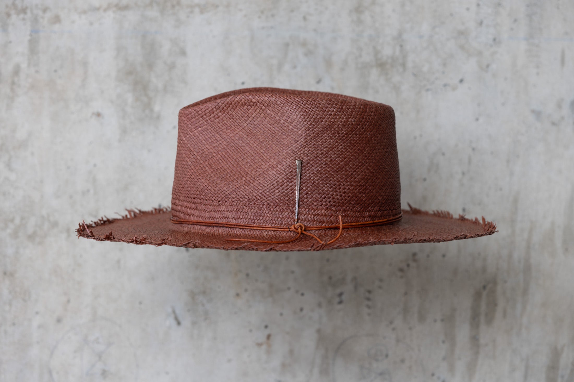 A chocolate brown straw hat with a teardrop crown and a frayed brim edge, with a leather cord wrap left side