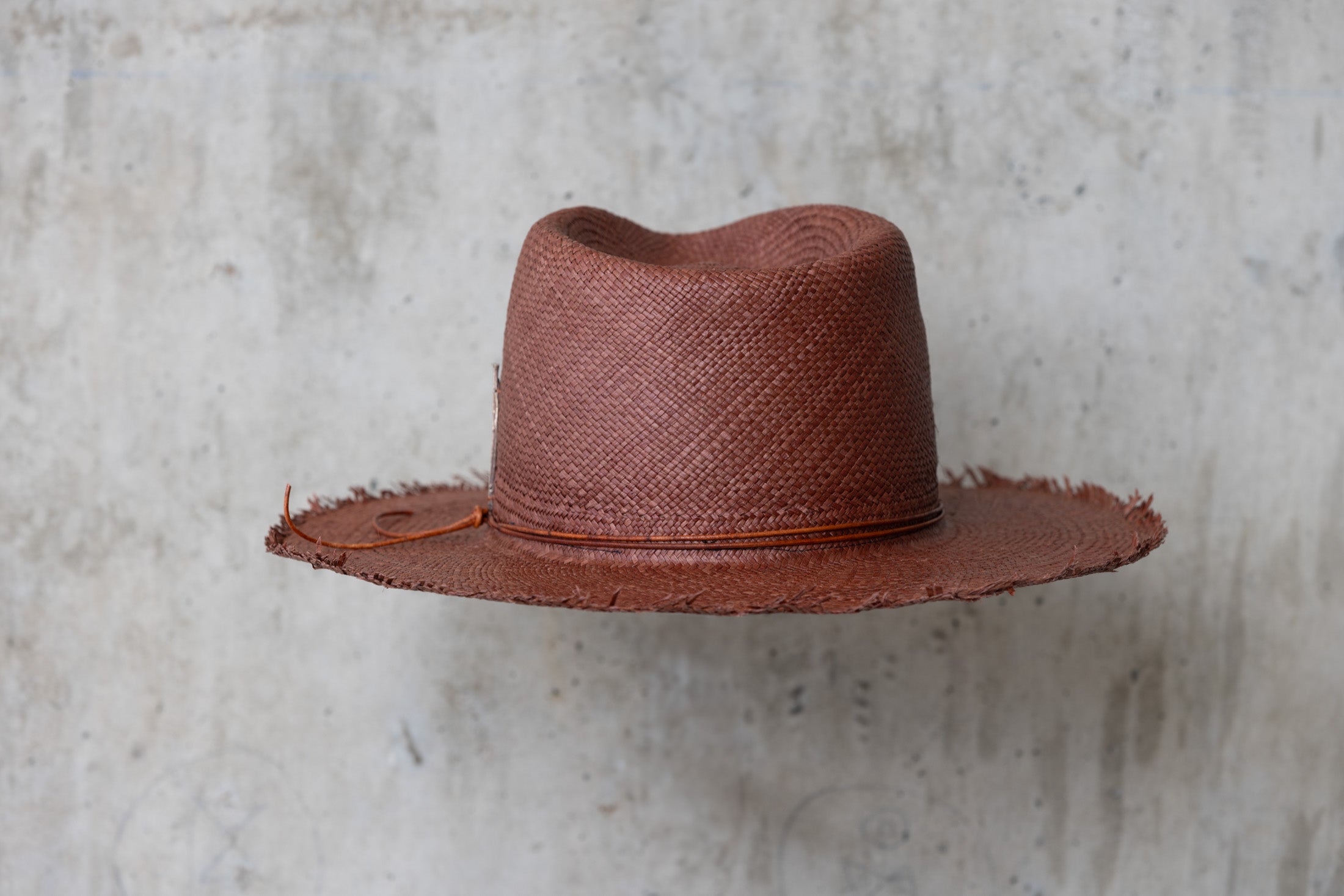 A chocolate brown straw hat with a teardrop crown and a frayed brim edge, with a leather cord wrap back side