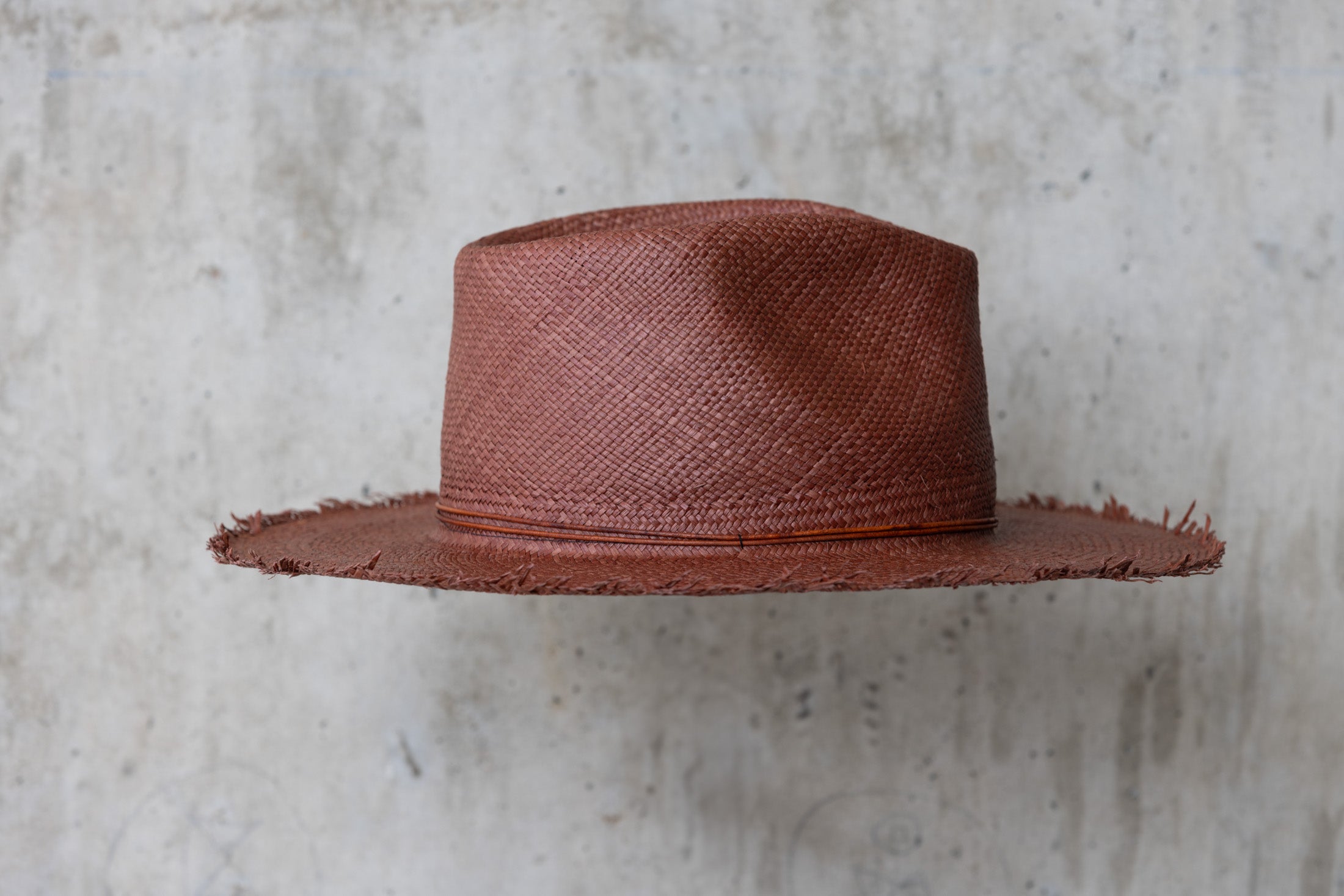 A chocolate brown straw hat with a teardrop crown and a frayed brim edge, with a leather cord wrap right side