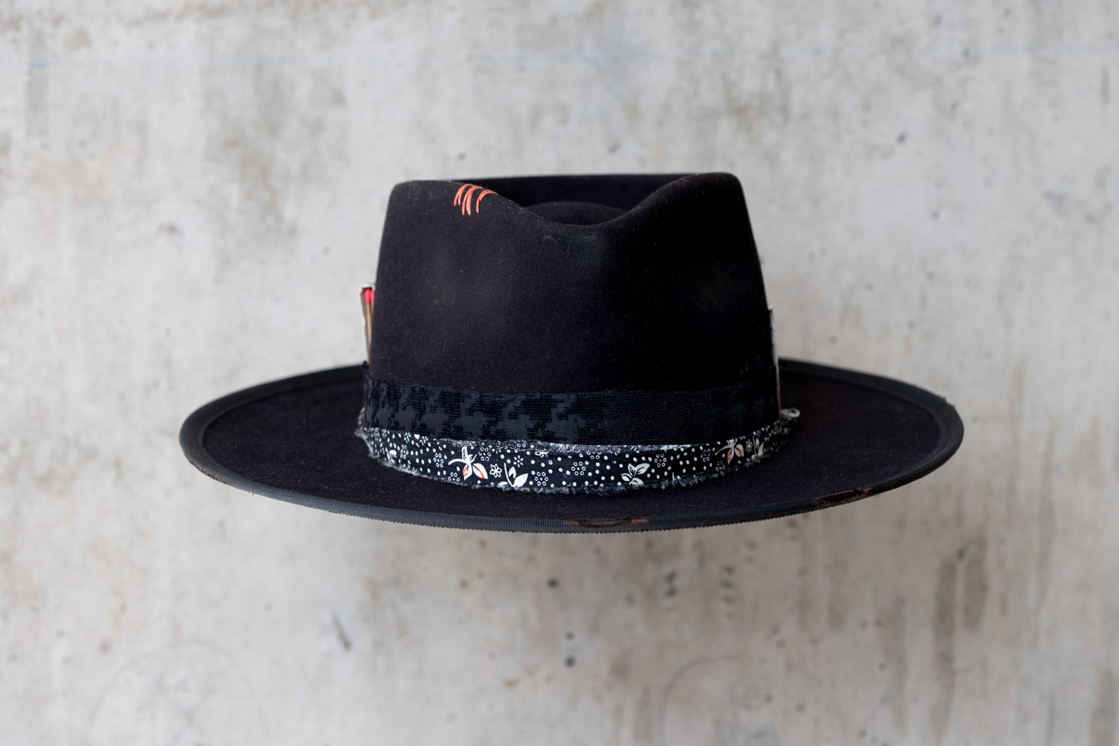A black fedora western beaver felt hat with a teardrop crown and a brim featuring pencil curls. It has a black houndstooth band with a floral wrap and faded red stitching for men and women front