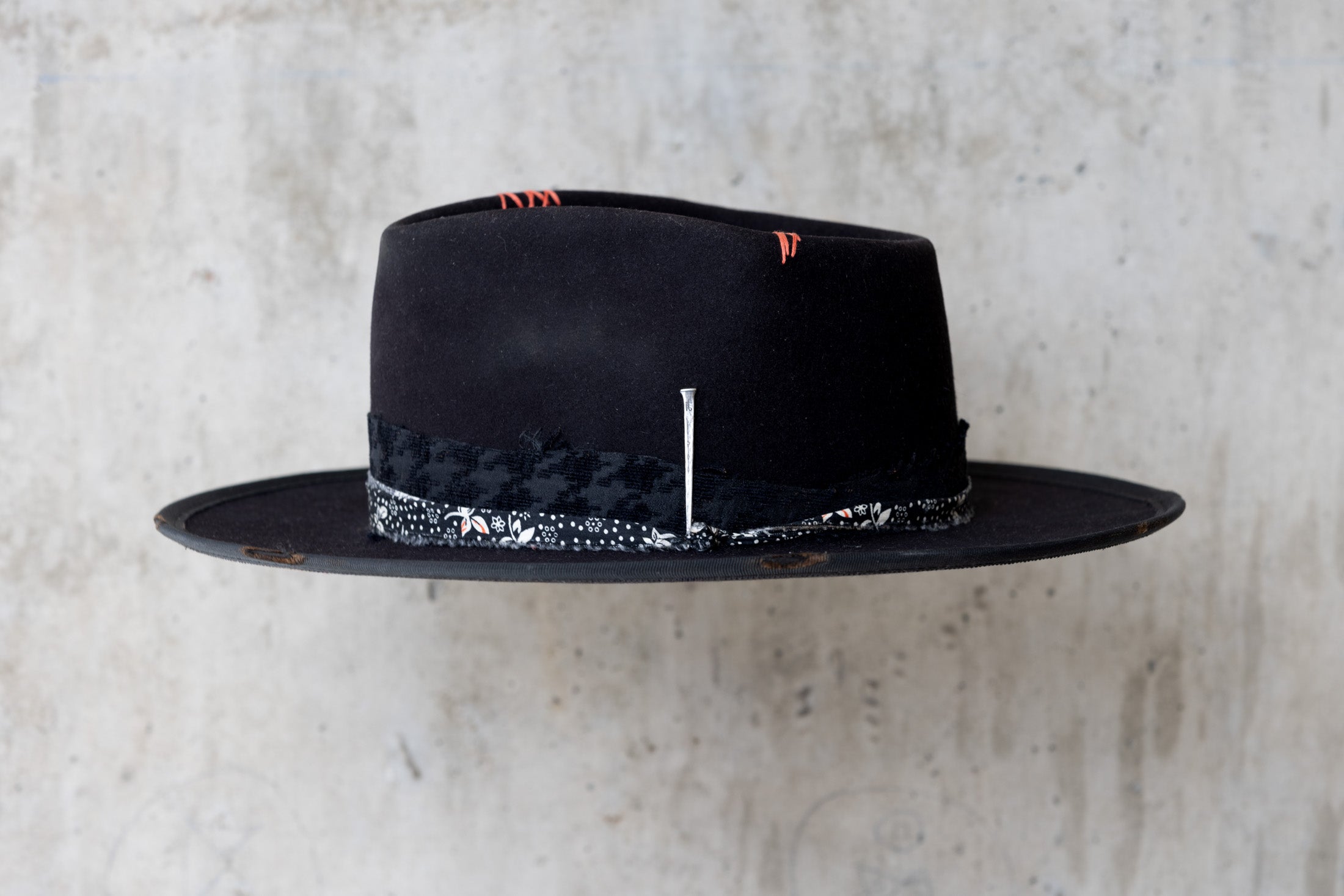 A black fedora western beaver felt hat with a teardrop crown and a brim featuring pencil curls. It has a black houndstooth band with a floral wrap and faded red stitching for men and women left side